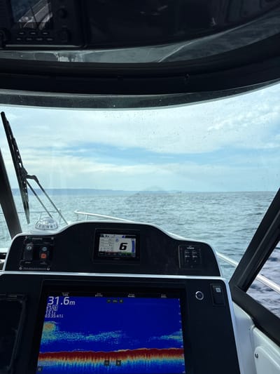 This image captures the interior of a boat cabin, focusing on the dashboard and windshield view. A speedometer displays a speed of "6," while a larger digital screen shows underwater sonar readings at a depth of "31.6m" with a time stamp. A windshield wiper sits at the base of a water-spotted windshield. The sky and water visible beyond the windshield suggest the boat is underway. Lenco switches are visible.