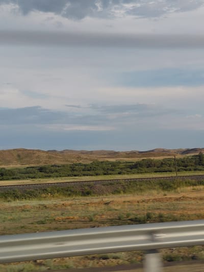 The image captures a landscape in Қапанбұлақ ауылдық округі, Kazakhstan, with a focus on the natural environment. In the foreground, a grey metal guardrail runs horizontally across the frame, suggesting a road or elevated viewpoint. Beyond the guardrail, a grassy plain with scattered vegetation stretches towards rolling hills in the mid-ground. These hills are covered in dry grass and sparse bushes. A line of green trees runs horizontally across the image, separating the grassy plain from the hills. Further back, a railway track is visible, running parallel to the line of trees and hills. The sky above is cloudy, with a mix of grey and lighter blue hues, indicating an overcast or partly cloudy day. The overall scene depicts a rural or semi-arid environment with a dominant presence of natural elements.