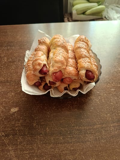 A close-up, eye-level shot displays a plate of seven pigs-in-a-blanket resting on a white paper napkin. The pigs-in-a-blanket are arranged in a pile within a dark, scalloped-edge bowl. Each pastry is golden brown with a light dusting of sesame seeds and features a visible segment of a reddish-brown sausage. The pastries appear to be made from a flaky dough wrapped around the sausages in a spiral pattern. The bowl sits on a dark, textured wooden surface, possibly a table. In the background, out of focus, is a metallic surface and a dark, indistinct object. To the upper right, a clear plastic container holds several light green, elongated items, possibly cucumbers, in front of a white plastic bag. The lighting is bright, creating some glare on the metallic surface. The overall scene suggests a casual indoor setting, likely a kitchen or dining area. The image does not contain any specific identifying markers that would place it within the city of Novoselytsia, Ukraine.
