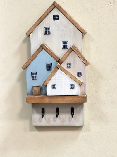 This is a close-up, eye-level, vertical shot of a wooden decorative wall hanging shaped like a cluster of houses. The houses are painted in a distressed white and pale blue, with natural wood trim around the roofs and window frames. Each house has small, dark blue windows with visible panes. The houses are layered on top of each other, with the largest at the top and progressively smaller ones below. A small, light brown wooden door is visible on the lowest blue house. Below the houses is a horizontal wooden shelf, and beneath that, the main body of the wall hanging features three black metal hooks for keys. The background is a textured, light yellow wall. There are no people or animals present. The object is mounted on a wall, casting subtle shadows. The image does not contain any specific visual information that would allow for its location to be identified as Maloyaroslavets, Russia.