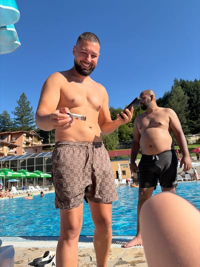 The image depicts an outdoor scene at a swimming pool, likely located at a resort in Chiflik, Bulgaria, on a sunny day. In the foreground, a man with a beard and short hair is smiling and holding a small cylindrical object in his hand. He is wearing patterned swim shorts. To his right, another man, also shirtless and with a beard, is holding a black electronic device to his ear and looking to the side. He is wearing black swim shorts. The background shows a bright blue swimming pool with people swimming and relaxing. Further back, there are buildings with balconies, green umbrellas, and trees, characteristic of a mountainous resort area. The overall atmosphere suggests a leisurely day at a vacation destination.