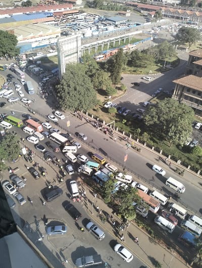 An aerial view shows a busy urban street scene with heavy vehicular and pedestrian traffic. The street is lined with cars, vans, and buses. A large bus depot or terminal is visible in the background with numerous parked buses. A green space with trees separates the depot from the street. Buildings are interspersed throughout the image, indicating a mixed-use area. The presence of pedestrians suggests a vibrant and bustling city center.