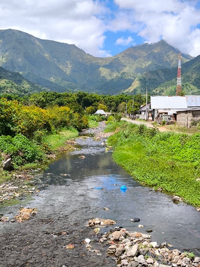 The image captures a polluted river running through the village of Sembalun Bumbung, Indonesia, set against a backdrop of mountains. The riverbed is littered with rocks and debris, including plastic bags and other forms of trash. The water appears murky and stagnant. Lush green vegetation lines both banks of the river, contrasting sharply with the pollution within it. A road runs along the river's right bank, flanked by simple buildings with corrugated metal roofs. A red and white telecommunications tower rises in the distance. The sky is partly cloudy.