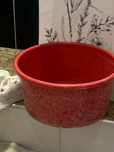 In Glodeni, Moldova, a red plastic bucket with a textured exterior sits on a white surface, possibly a countertop or bathtub ledge. A white athletic shoe with gray accents is visible to the left of the bucket. Behind the bucket, a wall features a black tiled section on the left and a white paper with a black and white botanical drawing on the right. The floor appears to be tiled with a stone-like pattern. The lighting suggests an indoor setting, possibly a bathroom or utility area.