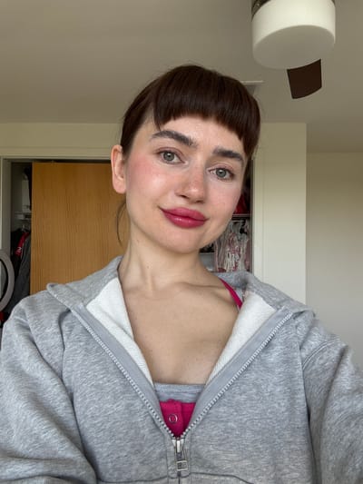 An indoor portrait captures a young person, likely a woman, from a slightly high angle, rotated 90 degrees counter-clockwise. She has fair skin with visible freckles on her nose and cheeks, dark brown hair cut with short, straight bangs, and striking green eyes. Her lips are adorned with a reddish-pink lipstick, and a soft blush is applied to her cheeks. She looks directly at the camera with a gentle, inviting smile.
She is wearing a casual grey hooded zip-up sweatshirt with white lining visible inside the hood and along the zipper. A bright pink top or bra strap is peeking out from underneath the partially unzipped sweatshirt.
The background suggests an interior setting, possibly a bedroom or personal space in Springfield, United States of America. Above her head, a white cylindrical light fixture with a dark brown, curved element is visible, resembling a modern ceiling fan or light fixture. Behind her right shoulder, a wooden wardrobe or closet door is partially open, revealing a glimpse of folded clothes or items inside. Towards the bottom of the frame, a segment of a circular ring light is just visible, suggesting the image might be a selfie or taken with supplementary lighting. The lighting appears soft and natural, indicative of daytime. No text is visible in the image.