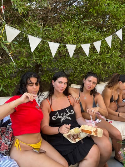 The image depicts an outdoor social gathering of four adult females seated on a light-colored sofa in Binyamina - Givat Ada, Israel. The background is composed of dense green foliage, with a string of white triangular pennant flags and yellow and white streamers indicating a celebratory atmosphere.
From left to right:
1. A woman with dark, curly hair wears a red short-sleeved top and yellow string bikini bottoms. A smartphone rests on her right thigh. She has food residue on her upper lip and holds a small object, possibly a utensil or food, near her mouth with her left hand.
2. A woman with dark, straight hair wears a black sundress with orange straps and white embroidery, a gold-colored choker, and hoop earrings. She holds two rectangular, light-colored plates containing slices of cake—one chocolate, one a lighter color—and a white plastic fork in her right hand, gesturing towards the food. She looks directly at the camera.
3. A woman with dark hair pulled back wears a dark grey bikini top, multiple rings, and a pendant necklace. Her left hand holds a white plastic fork over a red and white striped fabric on her lap. Her right arm rests on the back of the sofa, touching the shoulder of the woman to her right. She looks directly at the camera.
4. A fourth woman, partially visible on the far right, has dark hair pulled back and wears a black bikini top and bottoms. She appears to be interacting with something off-frame.
The scene is illuminated by daylight, suggesting an daytime event.