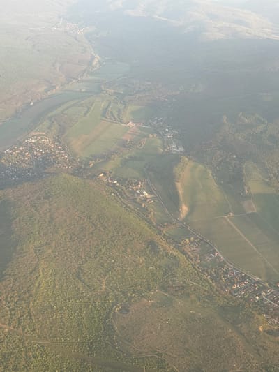 The image shows an aerial view of an area in Vecsés, Hungary, characterized by a mix of natural and developed landscapes. Fields with distinct boundaries are visible, interspersed with dense forested areas. A road lined with residential structures runs along a boundary between the fields and forests. A cluster of buildings is seen within the fields. The left side of the image is obscured by a haze.