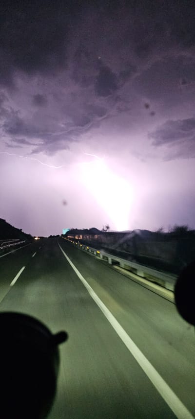 The image shows a highway at night with a thunderstorm in the distance. Lightning flashes illuminate the sky, casting a purple glow on the clouds. The highway is empty except for the perspective of the viewer, suggesting they are driving. The road is dark asphalt with white lane markings. Guardrails line the sides of the road, and some lights are visible in the distance, possibly from other vehicles or buildings. The background features dark hills or mountains. There is no visible indication of the city of Plasencia, Spain. The image appears to be taken from inside a vehicle.
