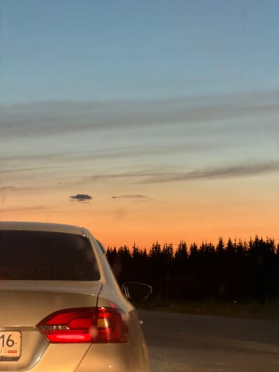 The image depicts the rear of a silver car, likely a sedan, parked on a paved road at dusk. The car's taillights are illuminated. A license plate with Cyrillic lettering and the number "16" is visible. The background features a silhouette of a dense forest against a sky colored with a gradient of pale blue at the top transitioning to a vibrant orange and yellow sunset closer to the horizon. A small, dark cloud is present in the sky above the tree line. The overall scene suggests a rural or suburban setting, possibly on the outskirts of Derzhavino, Russia, given the context. There are no people visible in the image.