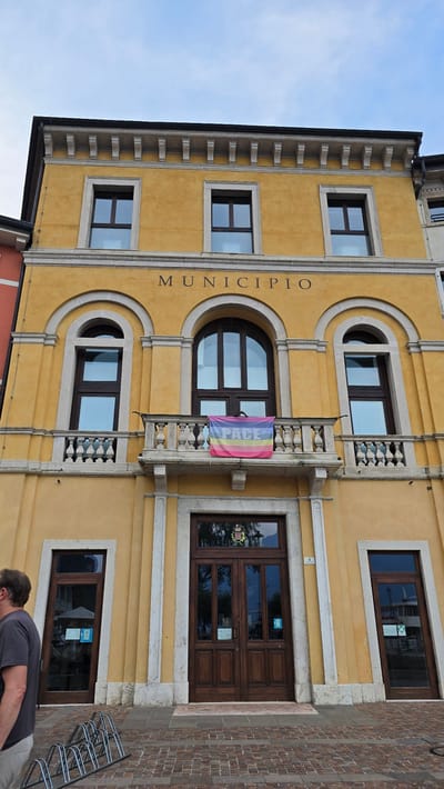 The image shows the facade of the Palazzo Pretorio, the town hall of Riva del Garda, Italy. The building is painted in a warm yellow color and features classical architectural elements, including arched windows, decorative moldings, and a prominent balcony. A banner with the word "PACE" (peace in Italian) and rainbow stripes is displayed on the balcony. The sky is overcast. In the foreground, a person in a grey t-shirt is visible on the left, and a bicycle rack is present on the paved ground. The entrance to the building consists of two large wooden doors.