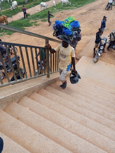 In this medium shot, a man walks up a concrete staircase carrying a large load of coiled ropes and other items balanced on his head. He is wearing a yellow t-shirt with blue trim and light-colored pants. In his right hand, he carries a black bucket. The man is on an outdoor staircase, likely in a public area. The background shows a sandy ground with some vegetation and cattle grazing. A metal railing runs alongside the staircase. In the distance, several motorcycles are parked, and other people can be seen. One man in a blue shirt and black shorts is standing near a motorcycle, looking at his phone. Another man in a black hoodie is sitting on a motorcycle. The overall scene appears to be a busy marketplace or a bustling area in Sauka, Nigeria. The time of day seems to be daytime, with bright lighting. The weather appears to be dry and sunny. The man's expression is neutral. The yellow t-shirt has the text "SUPER" and "TEA" printed on it.