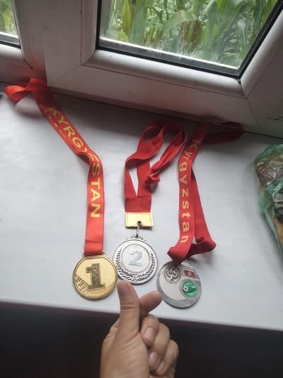 Three medals with red ribbons are laid out on a white surface, likely a windowsill, in front of a window. The medals are of different types and appear to be for first, second, and third place, although only the numbers "1" and "2" are clearly visible. The ribbons are inscribed with the word "KYRGYZSTAN" in yellow letters. A hand is visible in the foreground, with a thumb extended towards the medals. The background shows green foliage through the window, suggesting an outdoor environment. The medals are positioned on a white, flat surface that appears to be a windowsill. The lighting is natural, coming from the window. The scene suggests a display of achievements or awards. The location is identified as Baş-Bulak, Kyrgyzstan.