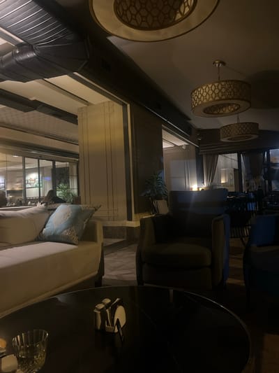 The image depicts the interior of a dimly lit, modern lounge or restaurant. In the foreground, a dark, glossy round table holds a glass of amber liquid, a salt and pepper shaker set, and a napkin holder. To the left, a cream-colored sofa with a light blue patterned throw pillow is visible. Behind the sofa, a dark armchair with a green hue is positioned. Further back, large glass windows reveal the exterior, with glimpses of other patrons seated at tables and interior lighting. The ceiling features industrial-style ductwork and two large, ornate pendant light fixtures with geometric patterns. The overall ambiance suggests an upscale, contemporary establishment. The location is identified as Bayrampaşa, Turkey, likely a commercial area within the city.