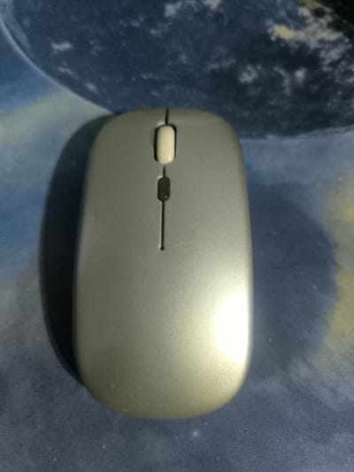 A silver wireless computer mouse with a white button on the top and a black scroll wheel in the middle, is laying on a blue and white patterned fabric surface. The mouse is laying on its back. The coordinates provided (5.9 latitude, 5.623 longitude) are located in the city of Sapele, Nigeria. The image was captured indoors. There are no people or other objects in the image.