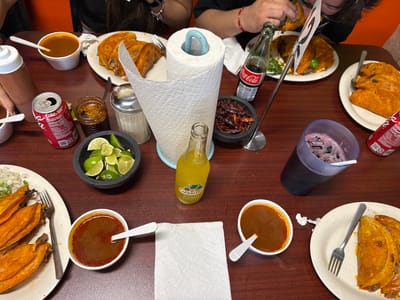 A high-angle shot depicts a table covered with food, condiments, and drinks typical of a casual Mexican restaurant setting. The table, appearing to be made of wood, is covered with plates of what appears to be tacos, accompanied by small containers of sauce. Multiple individuals are seated around the table, though their faces are mostly obscured, indicating they are in the process of eating or preparing to eat.
Condiments such as lime wedges, dried chilies, and sauces are scattered across the table. A paper towel roll is centrally placed for easy access. Beverages present include Coca-Cola in both bottles and cans, and a Jarritos pineapple soda. A dark-colored beverage is in a large plastic cup.
Based on the arrangement and type of food, this scene is likely to be at a birria taco restaurant. Birria tacos are popular in Chicago, United States, so this scene could plausibly be located in that city.