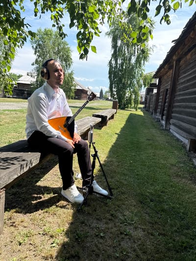 An individual wearing headphones and a white collared shirt sits on a wooden bench holding a balalaika. They are positioned outdoors, with a microphone on a stand in front of them. Behind the individual, a row of traditional wooden buildings forms a backdrop, suggesting a historical or ethnographic setting. The environment includes a grassy area with trees and a bright, partly cloudy sky. This scene appears to be within the territory of the Shushenskoye Museum-Reserve, a place known for its preserved historical buildings from the late 19th and early 20th centuries, showcasing Siberian rural life.