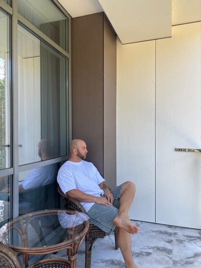 A bald man with a beard wearing a white t-shirt and grey shorts is sitting on a wicker chair on a balcony. He is looking to his right with his legs crossed. A glass-topped table with a wicker base is in front of him. The balcony has a marble-tiled floor and is enclosed by large windows on the left and paneling on the right. The man's reflection is visible in the window. The scene is set in an outdoor area with trees visible in the background through the window. The man is positioned in the center-right of the frame.