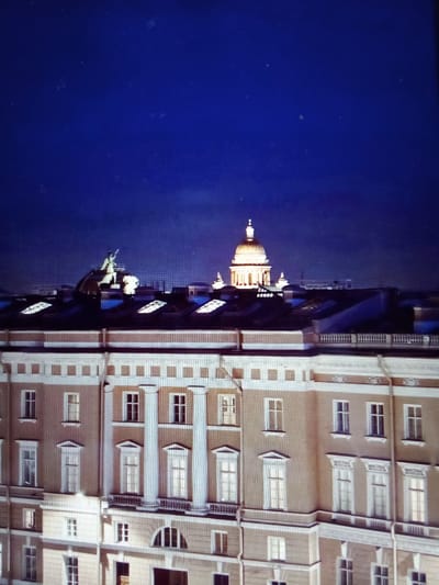 The image shows a nighttime view of a city skyline dominated by classical architecture. In the foreground, a multi-story building with a light-colored facade features numerous windows and ornate columns. The roof of this building has several skylights. Further in the background, a prominent golden dome, illuminated from within, rises above other structures. To the left of the dome, a statue on horseback is visible atop another dome. The sky is a deep, dark blue, suggesting it is late evening or early morning. The presence of illuminated domes and statues, along with the classical architectural style of the buildings, strongly suggests a location like Saint Petersburg, Russia, known for its grand palaces and cathedrals. The scene does not contain any individuals or direct human activity. The location "посёлок дачного хозяйства «Архангельское»" is a dacha settlement, which is typically characterized by residential houses and private gardens, and does not fit the architectural style and scale depicted in the image. Therefore, the provided location is inconsistent with the visual evidence.