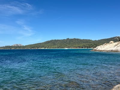 This image depicts a coastal scene near Hyères, France, likely on the Île de Porquerolles or the Giens peninsula. The foreground is dominated by clear, turquoise water with visible rocks and pebbles beneath the surface. The water exhibits gentle ripples, suggesting calm conditions.
In the middle ground, a vast expanse of deep blue sea stretches towards the horizon. Two sailboats are visible on the water, one a white catamaran and the other a white monohull, both appearing to be stationary or moving slowly.
The background features a densely forested, green hillside that slopes down to meet the sea. On the far left of the image, a rocky, mountainous outcrop juts out into the water. The sky is a vibrant, clear blue with a few wispy white clouds and a faint contrail. The overall impression is one of a serene, natural coastal landscape under a sunny sky. There are no people or visible structures apart from the sailboats.