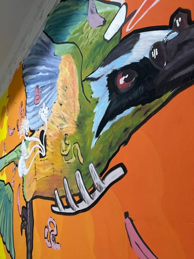 A vibrant, stylized mural is depicted on an orange wall in Santa Marta, Colombia. The artwork features a large, close-up depiction of a bird's head and wing. The bird has striking red eyes, a black beak with white and light blue accents, and a black and white striped head. Its wings are a mix of green, blue, and yellow feathers.

Scattered around the bird are various abstract and figurative elements painted in bright colors. To the left of the bird, there are two white, elongated figures with dog-like heads and wispy white appendages, possibly representing spirits or abstract representations of life. A pink, stylized figure resembling a flamingo is also visible on the blue wing. Below these, green leaves extend from a dark brown branch.

To the right of the bird's head, a pink banana-like shape is painted on the orange background. Several other abstract pink and yellow shapes are scattered throughout the mural. The overall style is cartoonish and energetic, with bold black outlines defining the shapes. The orange wall serves as a stark backdrop, making the colorful imagery stand out. The lighting suggests it is daytime, with no shadows indicating direct sunlight.