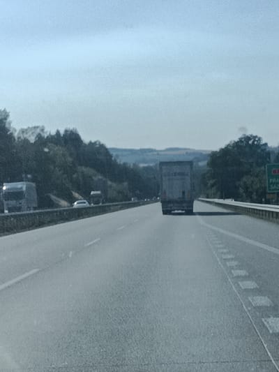 The image shows a highway scene, likely in Divišov, Czechia, on a clear day with bright sunlight. The perspective is from a vehicle traveling on the highway, looking forward. The foreground is dominated by the asphalt road with white lane markings, including dashed lines and solid lines indicating the shoulder.
Several vehicles are visible on the highway. In the distance, a large gray truck with a rectangular trailer is traveling in the same direction. To the left, another truck, also gray, is seen with its headlights on, appearing to be in a different lane or slightly ahead. A white passenger car is positioned between these two trucks, also on the left side of the highway. Further down the road, other vehicles can be seen as small specks, suggesting ongoing traffic.
The sides of the highway are lined with dense green trees and vegetation, creating a natural backdrop. In the far distance, rolling hills under a pale blue sky can be observed. On the right side of the road, a green highway sign with white text is partially visible. The visible text reads "PRA" and "SAZ," which likely indicates a road destination.
The overall impression is one of travel and transit on a sunny day, with a clear view of the road and the surrounding natural landscape. The time of day appears to be daytime, given the bright sunlight casting shadows and illuminating the scene. The weather seems pleasant and clear, contributing to good visibility.