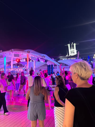 In an outdoor nighttime setting, a group of people are dancing on a wooden deck illuminated by vibrant pink and blue lights. A DJ is visible on a raised platform behind them, and a large cruise ship's superstructure is silhouetted against the dark sky in the background. The image does not depict Salobreña, Spain, or any recognizable urban elements.