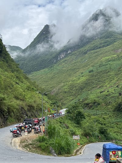 The image shows a steep, green, mountainous landscape, partly shrouded in clouds, in Yên Minh Commune, Vietnam. A narrow road winds along the edge of the mountain. Several parked motorbikes and a few standing people are visible on a roadside platform. A vendor's stand, covered with a blue tarp, is situated beside the road. The road surface is asphalt. Power lines and poles run along the hillside.