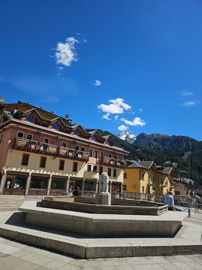 The image shows a sunny day in Ponte di Legno, Italy. A stone fountain is in the foreground, and behind it, a row of colorful buildings with wooden balconies lines a street. Mountains covered in trees rise in the background, with a snow-capped peak visible in the distance under a bright blue sky dotted with white clouds. A man in a blue shirt and jeans is sitting on the edge of the fountain, facing away from the camera. Another person, wearing a red shirt, is visible in the background near one of the buildings. The architecture suggests a European mountain town setting.