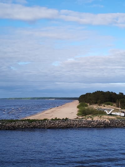 The image shows a view of the Volgo-Balt Canal named after V.I. Lenin in Ozernoe Ust'e, Russia. The foreground depicts dark blue water with gentle ripples. A rocky breakwater stretches horizontally across the middle ground. Beyond the breakwater is a sandy beach, bordered by a line of trees. A sign with the inscription "Волго-Балтом В.И. Ленина" is visible next to a boat-shaped structure. The sky above is partly cloudy with a mix of blue and gray.