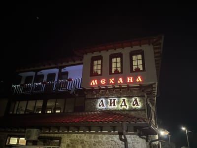 The image captures the exterior of a restaurant named "Механа АИДА" (Mehana AIDA) at night, located in Smilyan, Bulgaria. The building is constructed of stone on the lower levels and white plaster on the upper level. The restaurant's name is displayed in brightly lit Cyrillic letters above the entrance. The roof is tiled in reddish-brown hues. Windows are visible on both levels, some lit from within. A balcony with lights decorates the upper portion of the building. Streetlights illuminate the surrounding area.