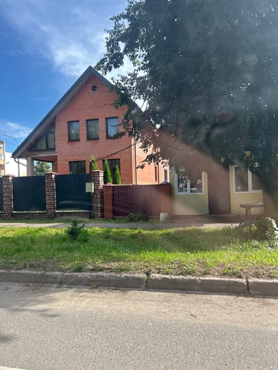 The image depicts a residential street scene in Lotoshino, Russia. In the foreground, a grey asphalt road is visible with a concrete curb separating it from a strip of green grass and some foliage. Behind the grass, a dark green metal fence runs horizontally, supported by brick pillars. To the left of the fence, a tall, multi-story brick house with a steep gabled roof and dark-framed windows is partially obscured by a large tree with dark green leaves. The house appears to be a modern structure with a balcony or covered patio on the upper level. To the right of the brick house, a smaller, single-story yellow house with white trim and windows is visible. A dark door is present on the yellow house. The sky above is blue with scattered white clouds. The overall impression is of a suburban or rural residential area.