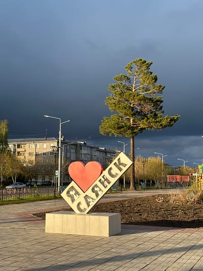 The image shows a view in Sayansk, Russia, with a prominent "I <3 SAYANSK" sign in the foreground. The sign features a red heart shape and Russian text. A tree stands adjacent to the sign, and behind it are multi-story apartment buildings and streetlights. The sky in the background is a mix of dark, stormy clouds and brighter areas. The foreground consists of a paved walkway. A small playground area can be seen further in the background to the right.