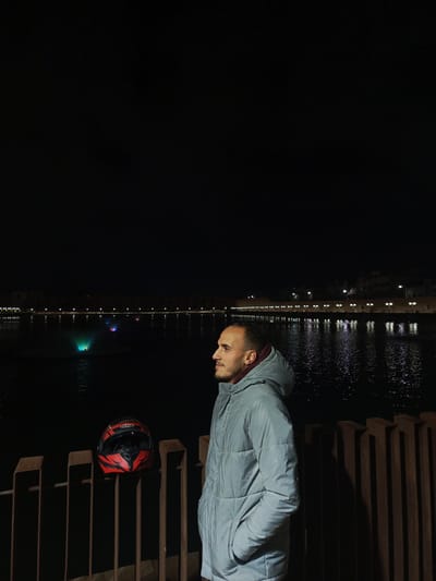 A man stands at night in Al Machouar – Stinia, Morocco, near a body of water with illuminated fountains and building lights reflecting on the water's surface. He wears a light-colored hooded jacket and is positioned near a fence with a red and black motorcycle helmet resting on it. The background is dark, with the lights from the buildings and fountains providing illumination.