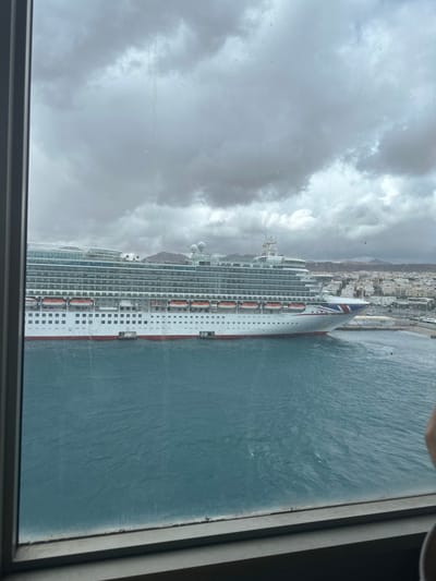 The image presents an exterior view through a window, showcasing the harbor of Puerto del Rosario, Spain, under an overcast sky. A large cruise ship, identified as "AZURA" by text on its bow, dominates the midground. The ship features a white hull with a red stripe along the waterline, blue-green balcony decks, and numerous orange lifeboats secured along its side. Its bow is oriented towards the right, displaying a Union Jack flag design. The harbor water is dark blue-green with visible surface ripples. To the right of the ship, a segment of the city of Puerto del Rosario is visible, comprising tightly packed light-colored buildings. Low, arid, reddish-brown hills rise in the distant background behind the cityscape. The sky is entirely covered by dense, grey clouds. The foreground includes a grey window frame, with minor smudges or water marks visible on the glass surface. No individuals are clearly discernible within the scene.