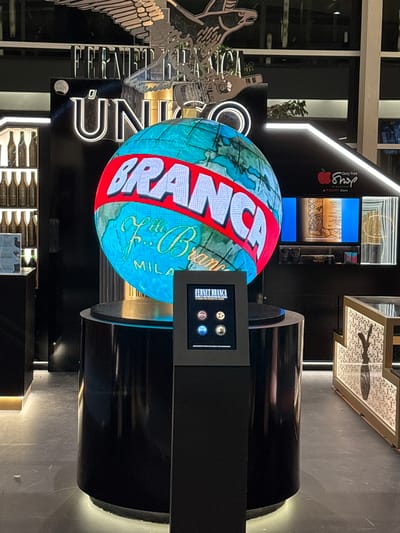 The image shows a retail display inside a duty-free shop at Aeropuerto Internacional Ezeiza, Argentina. The central focus is a large, illuminated globe displaying the word "BRANCA" in red lettering across its surface, superimposed on a map of the world. Below the globe is a black pedestal with a touchscreen display showing four circular icons and text that appears to be related to "FERNET BRANCA".

To the left of the globe, a white sign with the word "UNICO" is visible, above which is a security camera. Further to the left, on a white shelf, are several rows of small bottles. To the right of the globe, a screen displays an image of a bottle and some text. Behind the globe, a large metal sculpture of an eagle is mounted. The overall environment is dimly lit, with accent lighting highlighting the display elements and the floor. The shop appears to be modern and high-end, with clean lines and a dark color scheme. There are no people visible in the immediate vicinity of the display.