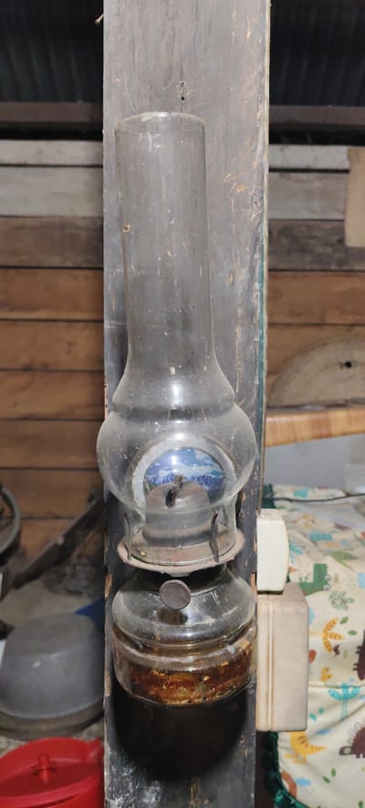 An antique oil lamp hangs on a weathered wooden post in an interior space in Jijiem, Indonesia. The lamp consists of a clear glass reservoir, a metal burner, and a glass chimney. The reservoir appears partially filled with a dark substance. The chimney is dusty and cylindrical. A small image of mountains is visible through the glass of the lamp. The wooden post is dark in color and shows signs of wear. Adjacent to the post is an off-white electrical outlet. In the background, there are weathered wooden planks and other household objects. A gray bowl and a red container are partially visible at the bottom left. A patterned fabric with dinosaurs is visible to the right of the post.