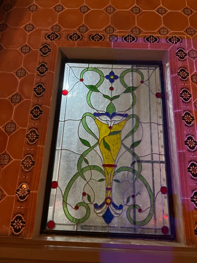 A stained-glass window is surrounded by terracotta-colored square tiles, some decorated with a stylized flower design, and detailed borders with alternating light and dark floral motifs. The window itself features a translucent background with decorative swirling patterns, overlaid with a design incorporating green vines, blue and yellow flower-like elements, and red circular accents. The window frame is black, and the whole assembly is set into a wall. The location is Santa Ana Yancuitlalpan (Chilhuacán), Mexico.