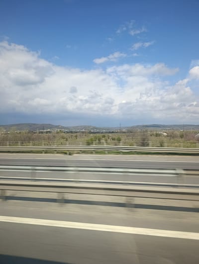 The image captures a scene in Ștefănești, Romania, as viewed from a moving vehicle on a highway. The foreground is dominated by the highway itself, with visible lanes marked by white lines and metal guardrails separating the road from the adjacent terrain.
Beyond the highway, the landscape features a relatively flat area covered with sparse vegetation, including small trees and grassy patches. In the distance, a chain of low hills or mountains is visible on the horizon, marked by what appears to be residential or urban development. Overhead, the sky is partly cloudy, with patches of blue visible between the clouds. The overall impression is one of a rural or semi-rural landscape seen in transit.