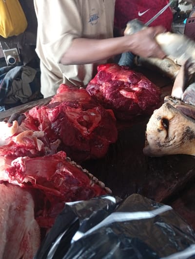 The scene depicts a butchery operation in Kofar Mazugal, Nigeria. A close-up view focuses on a dark, textured surface, likely a cutting board or table, covered with raw animal meat. In the center, two large, dark red cuts of muscle, one showing exposed bone, are prominent. To the left, a section of meat with a visible bone structure and red tissue is present. To the far right, a pale, irregularly shaped object resembling an animal's head or limb portion, with a rough texture and dark markings, rests on the table.
Above the meat, an individual wearing a light tan short-sleeved shirt with a blue logo on the left chest uses both hands to manipulate another pale, irregularly shaped piece of meat or carcass part. A metal knife is visible near their right hand, suggesting a cutting or processing action. Behind this individual, the upper body of a second person, wearing a red garment with a white Nike swoosh logo on the right chest, is partially visible. The background is dimly lit and includes a yellow bag hanging in the upper-left, and a small, light-colored square object with a dark circular dial is visible behind the meat on the left. A reflective black plastic bag occupies the lower-right foreground.