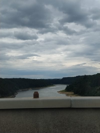 The image shows a wide river winding through a densely forested area. The sky is overcast with heavy, dark clouds. In the foreground, a light-colored concrete barrier runs horizontally across the bottom of the frame, with a darker, textured surface below it. A small, brown, hexagonal object is visible on top of the barrier, near the center of the image. The riverbanks are lined with dark green trees. In the distance, on the right side of the river, a sandy area is visible with a few small figures that appear to be people. The scene is consistent with a view from a bridge overlooking the Vranov Reservoir in Podolí I, Czechia.