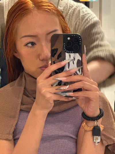 An individual with wet, reddish-orange hair is depicted indoors, likely within a hair salon in Toshima, Japan, though no specific street address or landmark is identifiable. The subject, with fair skin, occupies the central foreground, capturing a selfie using an iPhone with a black, patterned CASETiFY case. Their left eye, part of the nose, and mouth are visible, directed towards the phone screen. A brown towel is draped over their shoulders, covering a light purple ribbed top. The subject's hands exhibit long, pointed acrylic nails featuring clear designs with silver/white elements and rhinestones on the right ring finger. A black coiled wristband with a single metal key attached is visible on the right wrist, positioned above an orange scrunchie. Partially visible in the upper right background is the arm and shoulder of a second individual wearing a light-colored, textured garment, indicating the presence of another person, likely a stylist, attending to the subject's hair.