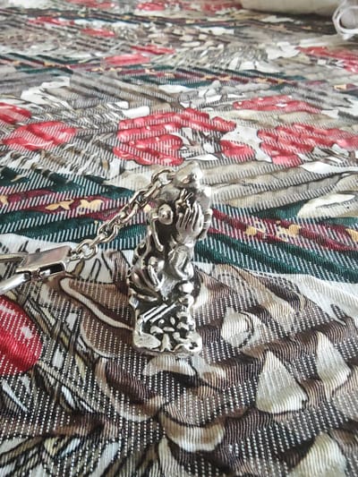 A silver colored keychain pendant in the shape of a kneeling human figure with hands clasped is shown from a high angle. The pendant is attached to a silver colored chain and a clasp. The pendant and chain are resting on a patterned fabric surface with red and green floral designs. The image was taken indoors on January 18, 2023, in Barntrup, Germany.