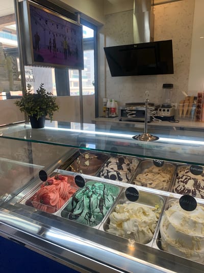 An indoor shot captures an ice cream display case filled with various flavors, including strawberry, mint, chocolate, vanilla, and what appears to be a coffee or caramel flavor, each labeled with small black signs. Above the display, a silver rack holds ice cream cones. In the background, a commercial blender and what looks like a crepe maker are visible on a counter. A flat-screen TV displays a group of people on a stage, possibly a performance. The scene suggests a dessert shop or cafe.