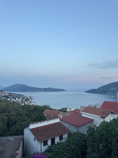 The image captures a serene panoramic view of a coastal town nestled beside a calm body of water, likely a bay or a lake, under a twilight sky. The scene is dominated by the soft hues of a setting or rising sun, casting a gentle glow across the water and the distant, hazy mountains that form the horizon.
In the foreground, several buildings with distinctive red-tiled roofs are visible, suggesting a residential or a small village area. Lush green trees surround these structures, adding to the natural beauty of the setting. The architecture of the buildings appears somewhat rustic, with white or light-colored walls.
The body of water is dotted with numerous small boats, indicating a leisurely activity or the presence of a harbor. The town itself is spread out along the coastline, with a density of buildings that gives it the character of a quaint European settlement. The overall atmosphere conveyed by the image is one of peace and tranquility, enhanced by the soft lighting and the vast expanse of water and sky.
Notable details include the clear division between the sky and the land, the muted colors of the twilight, and the numerous boats. The location cues, such as the mountainous backdrop and the Mediterranean-style architecture, point towards a European coastal region. The time of day is clearly indicated as twilight, with the sky transitioning between day and night. The weather appears calm and clear. No visible text is present in the image.