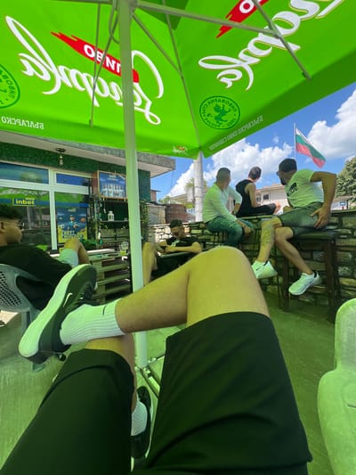 This image depicts an outdoor scene in Касапите, Bulgaria, on a clear, sunny day. The perspective is low-angle, from the viewpoint of a person seated under a large, bright green market umbrella, looking outwards.

The umbrella features mirrored white text, including "ОБОЛ" (Obol) and "ПРЕКРАСНО" (Prekrasno), as well as a green circular logo with a lion, the year "1803," and "СТАРА ЗАГОРА" (Stara Zagora) and "БЪЛГАРСКО" (Bulgarian) in Cyrillic.

In the immediate foreground, the legs and feet of the person taking the photo are visible, clad in black shorts, white ribbed socks with a black Nike swoosh, and black Nike sneakers with white soles.

To the left, another individual with dark skin, wearing sunglasses, a black t-shirt, and light-colored shorts, is seated on a grey plastic chair, looking towards the left. A small wooden table, constructed from pallets, holds a plastic cup containing a green beverage. Behind this person is a building with a facade covered in green artificial turf. The building features large windows displaying advertisements for "inbet," a betting company. One sign reads "ПУНКТ inbet," another "BET BUILDER," and a third shows images of footballers. A single light bulb hangs from the building's overhang. A white decorative lantern rests on a shelf to the right of the building's entrance.

In the mid-ground, slightly to the right of the pallet table, a male with dark hair, wearing a black t-shirt and dark shorts, is seated, looking down at a mobile phone.

Further to the right and in the background, three other males are seated at a stone wall or counter. The leftmost of these three wears a white long-sleeved shirt and blue jeans, looking away from the camera. The middle male wears a black tank top and black shorts, also looking away. The rightmost male wears a white t-shirt with "DUDE" printed on it, sunglasses, and grey shorts. He has a visible tattoo on his right calf and wears white Nike sneakers with air-cushioned soles. They are seated on high bar stools.

The background shows a blue sky with scattered white clouds. A Bulgarian flag, red, white, and green horizontally striped, waves on a pole against the sky, indicating the national context. Distant buildings and green foliage are also visible. The ground appears to be a light-colored paved surface.