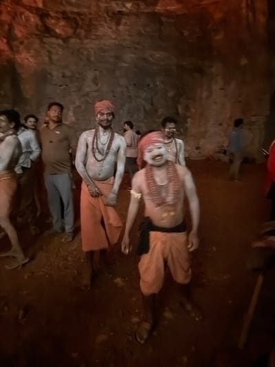 A group of people, some with their chests bared and adorned with necklaces, are gathered in a dimly lit, rocky cavern in Bethamcherla, India. The figures are mostly males, and some are wearing orange cloths around their waists. One individual in the foreground has white powder smeared on their face and chest, and a reddish-orange cloth wrapped around their head. The ground appears to be dirt and rock. The overall atmosphere is one of a ritualistic gathering or celebration.