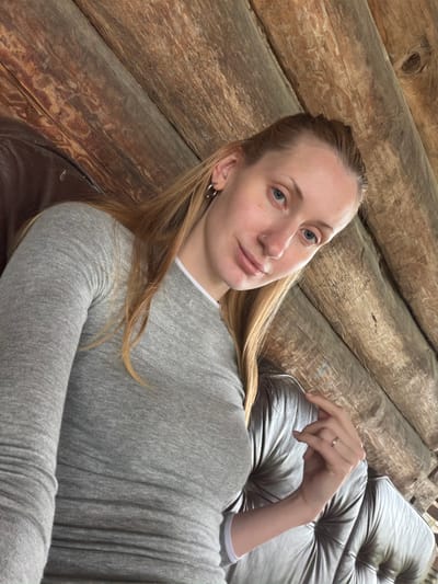 A young woman with blonde hair and blue eyes is pictured. She is wearing a grey long-sleeved shirt and a ring on her left hand. She is sitting on a tufted leather couch against a wooden wall. The location is not specified in Merkuriy, Russia.
