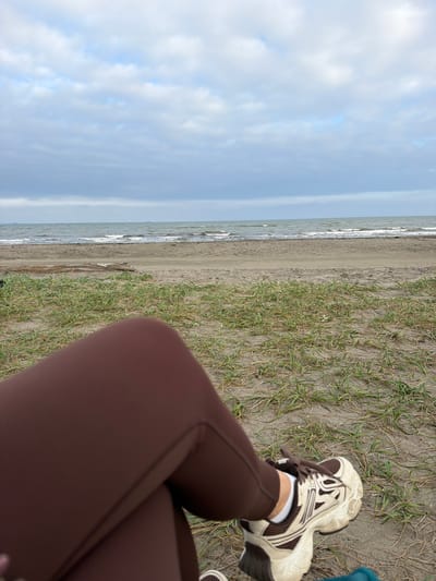 The image shows a person's legs and feet, wearing brown leggings and white and brown athletic shoes, crossed while sitting on a sandy beach. In the background, there is a calm sea with small waves under a cloudy sky. The beach has sparse patches of dry grass. The scene appears to be the coast of Sakhalin Island, potentially near Aniva, Russia, given the mention of the city.