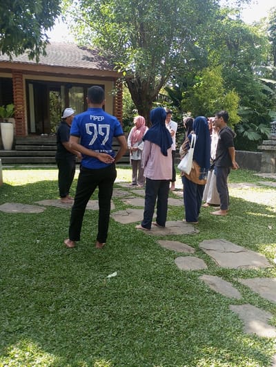 This image depicts an outdoor scene in Cibadak, Indonesia, under bright daylight. Approximately ten individuals are gathered on a manicured green lawn, which is traversed by irregular stone stepping paths.

In the foreground, a man stands with his back to the camera. He wears a blue t-shirt with "MPEP 99" prominently displayed on the back, along with additional text "JUNG BATU FE" below. He is clad in black pants and is barefoot, with his hands clasped behind his back. To his immediate left, another man, also barefoot, is partially visible, wearing a black shirt and a white head covering.

Further back and to the right, a group of approximately eight individuals is clustered. This group includes several women wearing hijabs of varying colors (e.g., pink, blue), and men in casual clothing, one of whom wears a black hoodie and glasses. Many members of this group also appear to be barefoot or wearing minimal footwear, standing on the grass or near the stone path. They are oriented towards each other, suggesting conversation or a shared focus.

In the background, a single-story residential building or villa is visible. It features a dark brown tiled roof, light brown walls, and large glass sliding doors or windows. Decorative vertical wooden slats are present on the left side of the building. A tall white planter with green foliage stands near the building's entrance steps. The entire scene is surrounded by dense, lush green vegetation, including large mature trees with full canopies and various shrubs, indicating a tropical environment. Shadows are present, consistent with an overhead sun.