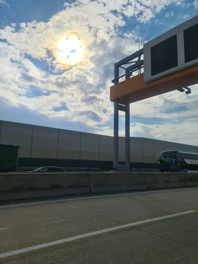 This image captures a scene on a highway, likely near Biedermannsdorf, Austria, under a partly cloudy sky with the sun visible and creating a halo effect. The foreground shows an asphalt road with white lane markings. To the left, a concrete barrier separates the road from a sound barrier wall. Beyond the sound barrier, a dark green trailer is partially visible, and a silver car is driving. A large overhead structure, possibly part of a toll booth or traffic information system, spans across the highway, supported by a concrete pillar. To the right of the pillar, a dark blue bus with white lettering and a circular logo is traveling on the highway. The bus appears to be a passenger transport vehicle, with "Burgenland" visible on its side. The overall environment suggests a travel or commuting scenario on a major road.